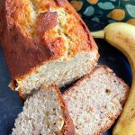 Banana bread loaf with two cut pieces to show the soft interior. It sits next to a banana and a green loaf tin decorated with pears and leaves.